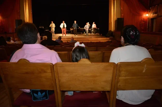 Zdjecie wnetrza teatru zrobione zza plecow widzow siedzacych w drewnianych fotelach