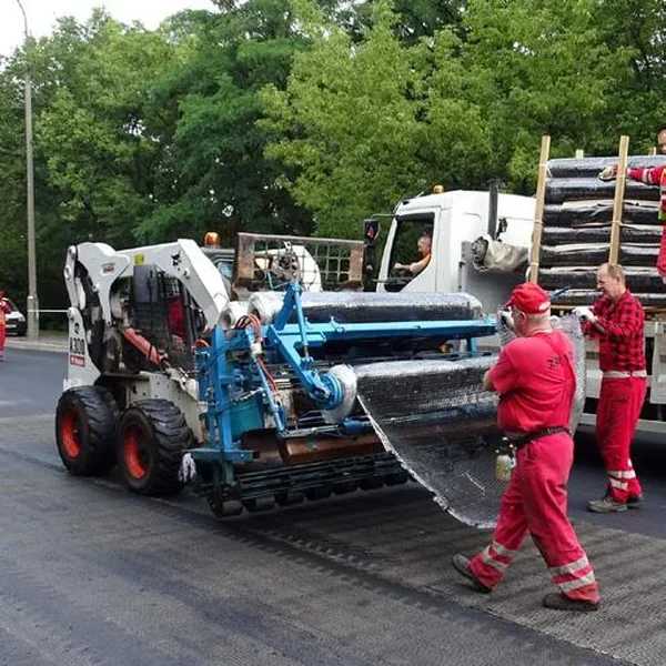 Zdjęcie maszyny na podwoziu gąsienicowym rozwijającej materiał w rolce na nawierzchni, obok pracownicy w czerwonych kombinezonach