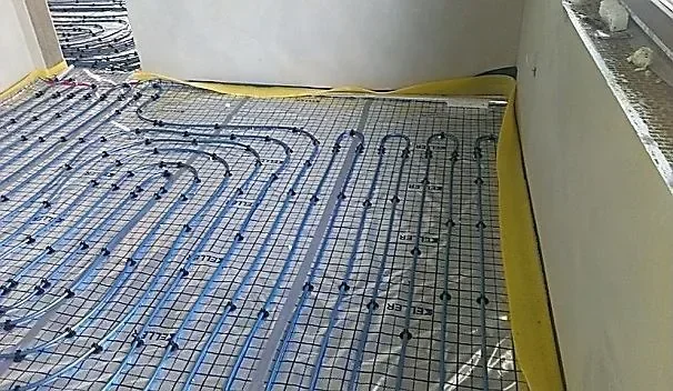 Niebieska rura ulozona wezykiem, z lukowymi nawrotami przy krawedziach pomieszczenia, przypieta spinkami do siatki na folii z zoltym pasem przysciennym